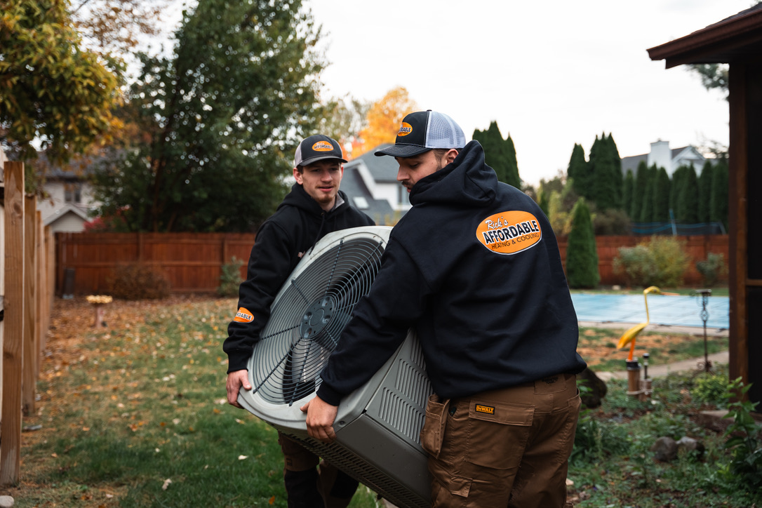 Two Rick's Affordable technicians carrying a new AC condenser unit to a customer's backyard